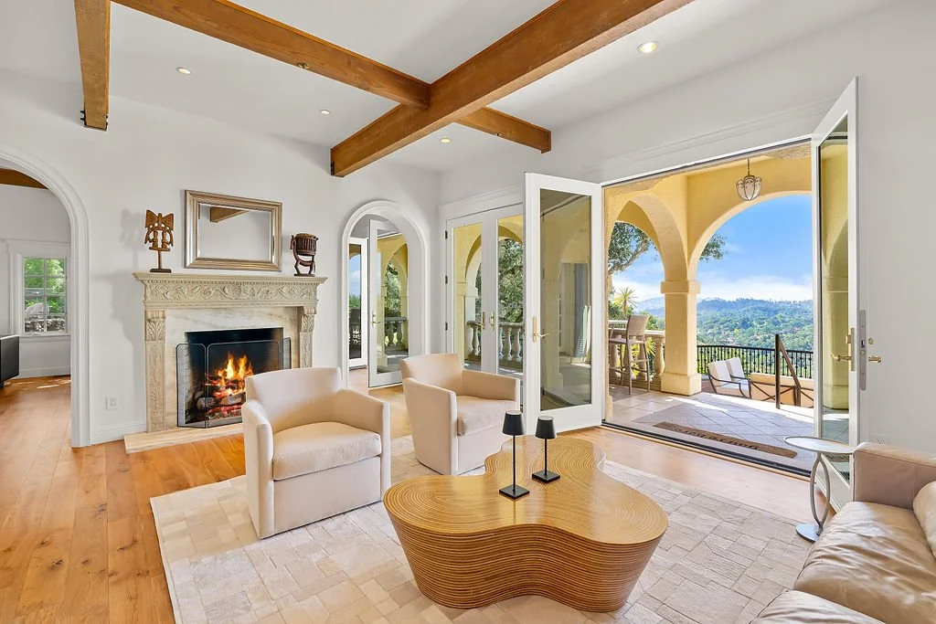 Living room with fireplace, beige armchairs, a wood coffee table, and large glass doors opening to a balcony with scenic mountain view.