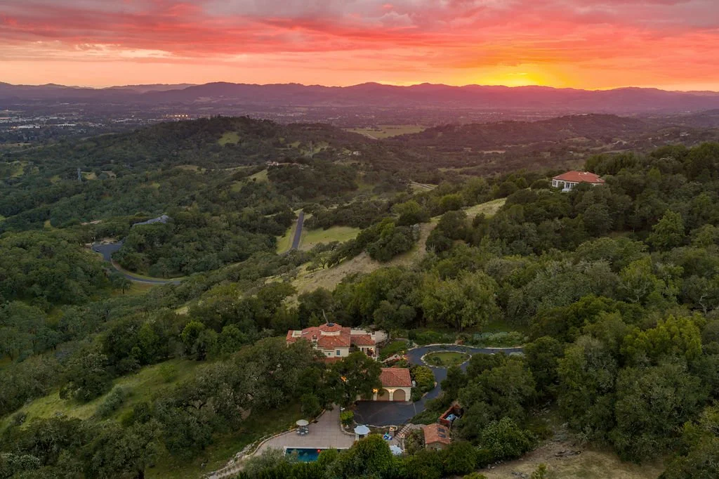Aerial view of a hilly landscape with houses and dense green trees at sunset, with colorful sky and distant mountains.