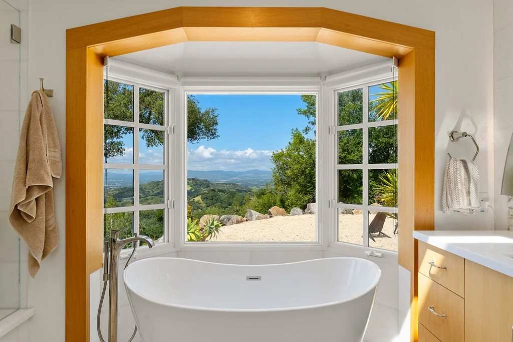 Freestanding white bathtub in a bright bathroom, overlooking a scenic landscape through a large bay window with multiple panes.