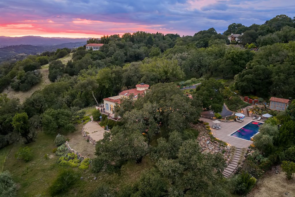 A scenic hillside residence with a swimming pool surrounded by lush trees and landscaped gardens at sunset.