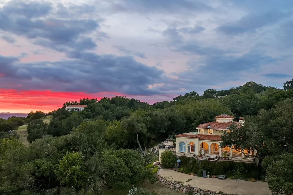 A large house with a terracotta roof and arched windows sits on a hillside surrounded by trees at sunset. A smaller building is visible further up the hill, with a colorful sky featuring pink, purple, and blue hues in the background.