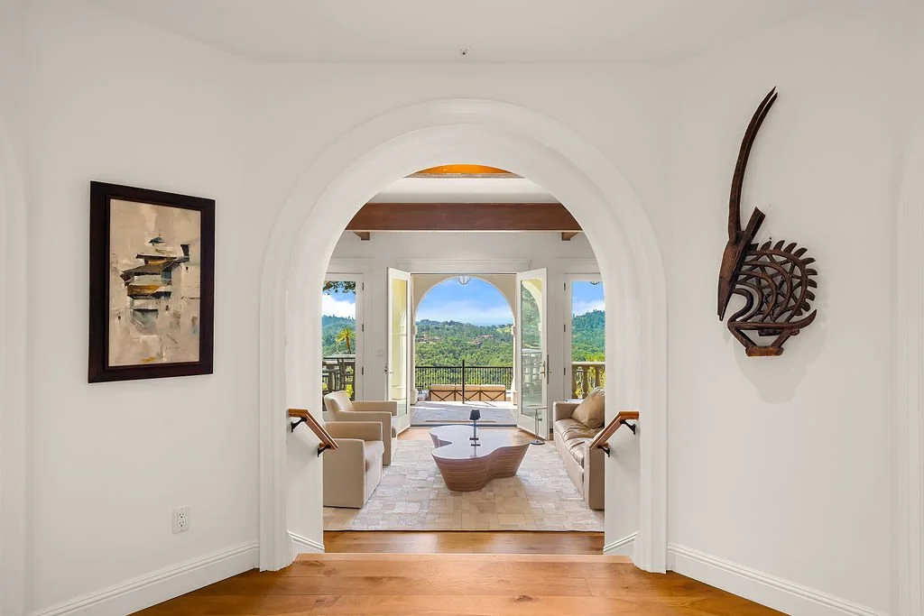 Living room with arched doorway, view of outdoor balcony, beige sofas, coffee table, artwork on walls, and scenic landscape viewing area.