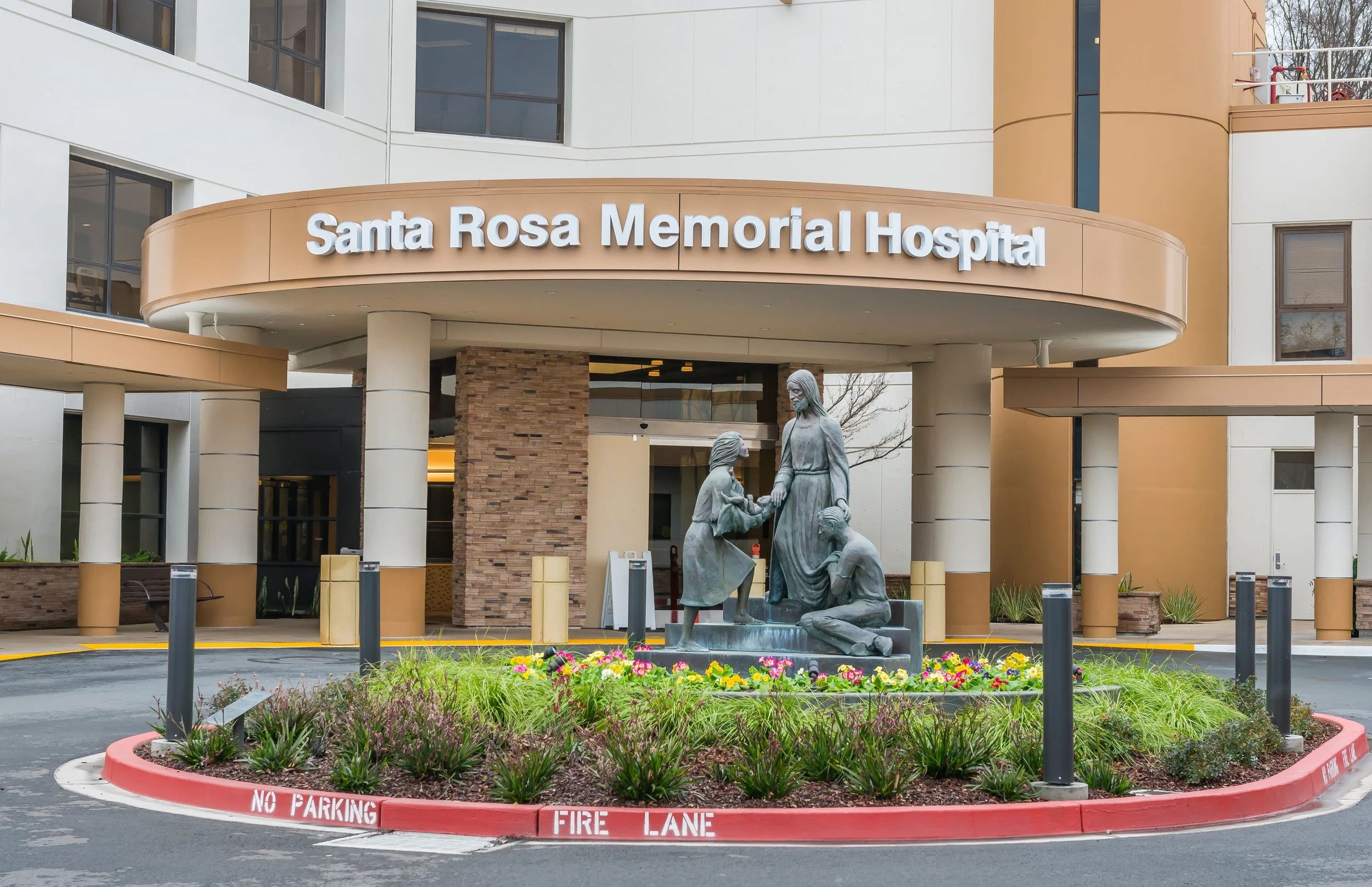 Exterior view of Santa Rosa Memorial Hospital entrance with a metal sculpture of a woman and children, flower bed, and no parking/fire lane signage.