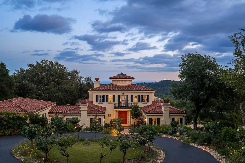 A large Mediterranean-style house with a red-tiled roof, surrounded by a lush garden and trees, under a partly cloudy sky at dusk.