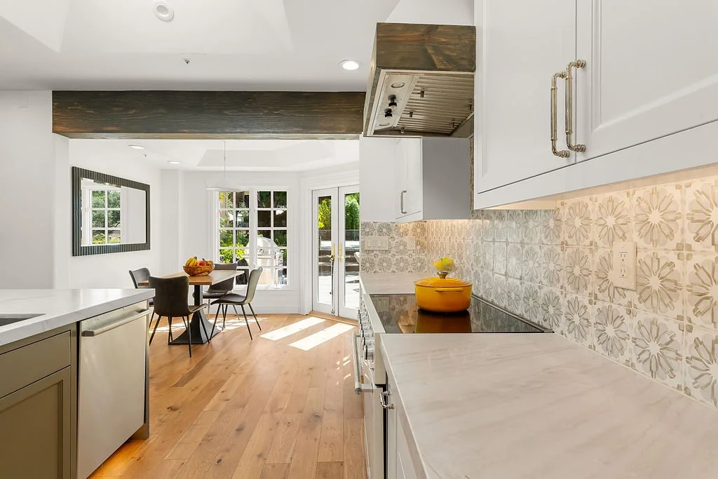 Bright kitchen with white cabinets, patterned tile backsplash, wooden flooring, and a dining area with a table and chairs near large windows and a door leading outside.