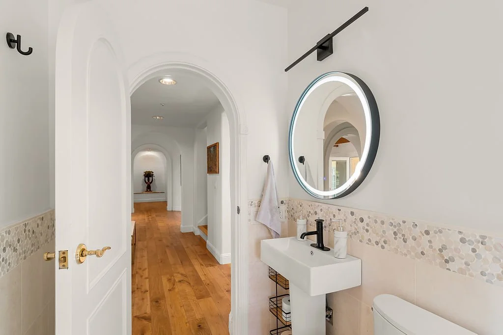 Bathroom with a small sink, round backlit mirror, white walls, a towel hanging on a black hook, and a toilet. Wooden flooring leads into a hallway with archways and a decorative vase with flowers in the background.