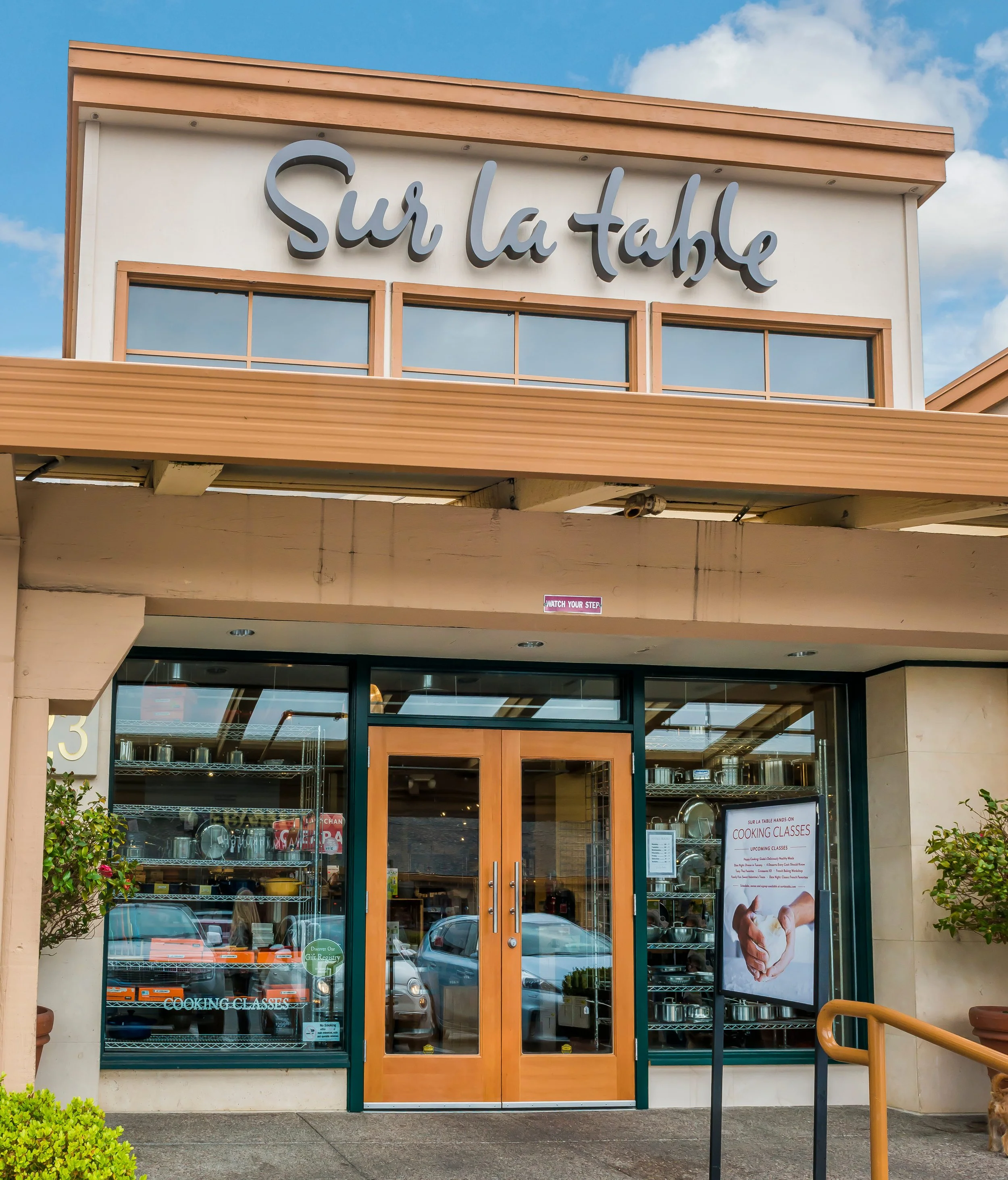 Exterior of a restaurant or store named Sur la Table with glass double doors, a sign for cooking classes, and a poster about upcoming classes, with some plants and cars visible outside.