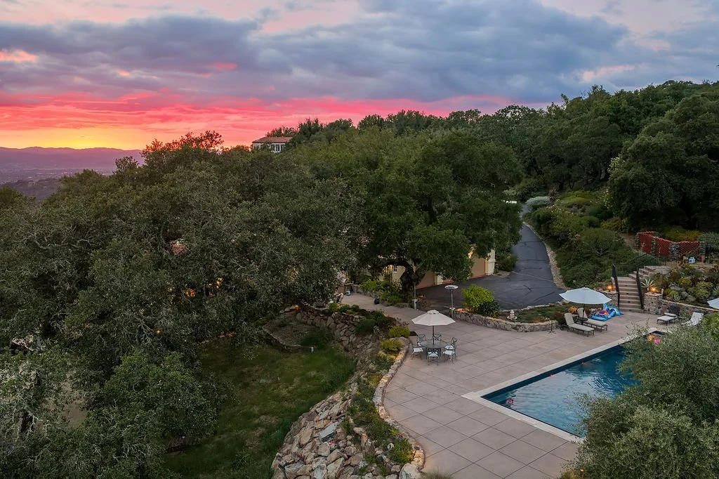 A backyard with a swimming pool, patio furniture with umbrellas, and surrounding trees during sunset or dusk.