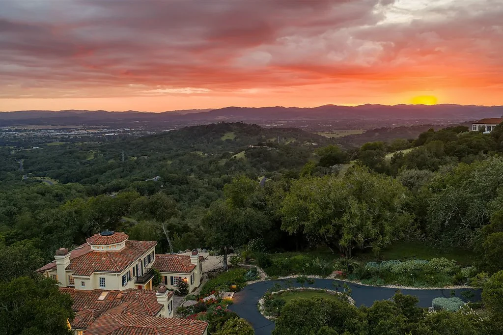 Sunset over a hilly landscape with a large house and a pool in the foreground, surrounded by trees and greenery.