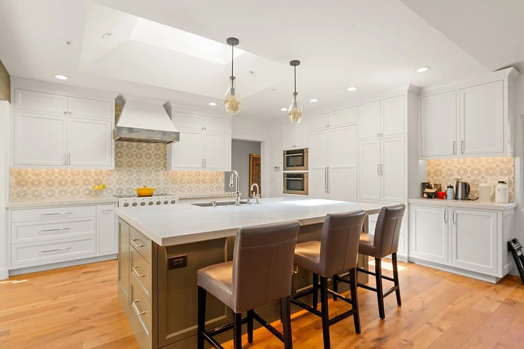 Modern white kitchen with an island, pendant lights, and built-in double ovens, featuring wood flooring and a patterned backsplash.