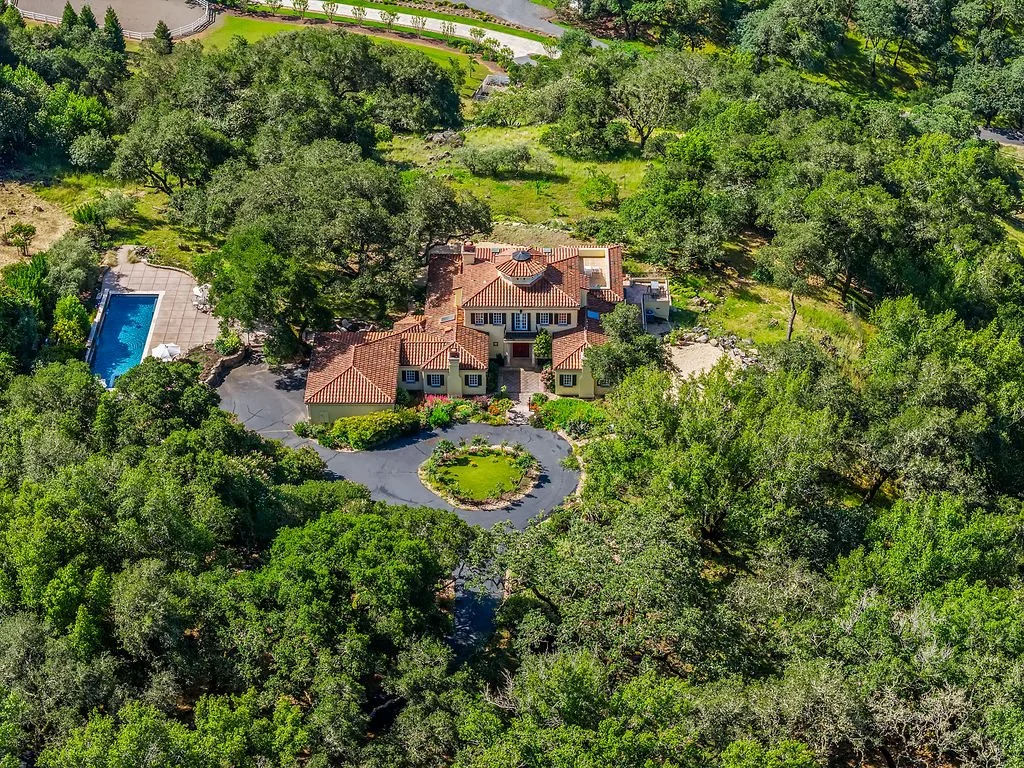A large house with a red-tiled roof, surrounded by trees and greenery, with a circular driveway leading to the entrance and a swimming pool on the left side.