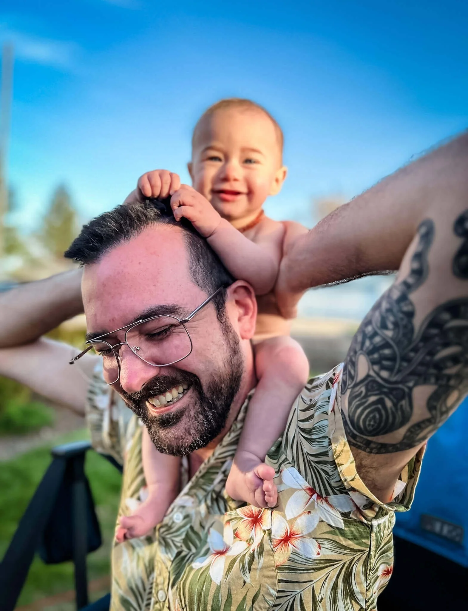 A smiling man with glasses and a beard carries a baby on his shoulders outdoors during daylight.
