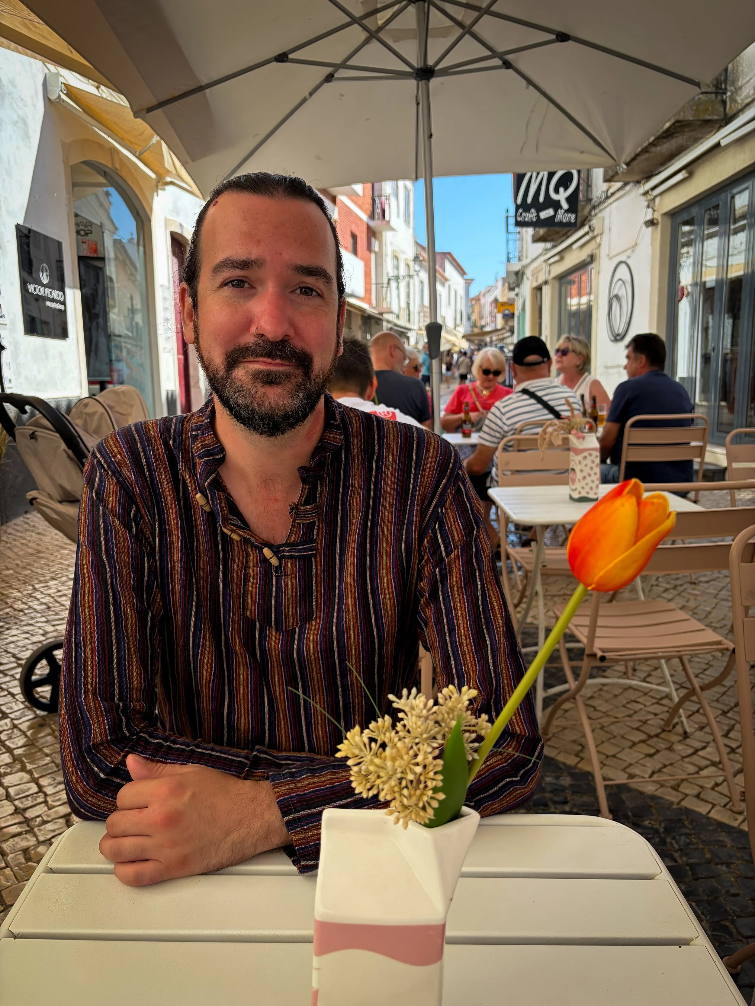 A man with dark hair and a beard sitting at an outdoor cafe table beneath a large umbrella, with a background of people dining and walking along a cobblestone street, and a vase of flowers on the table.