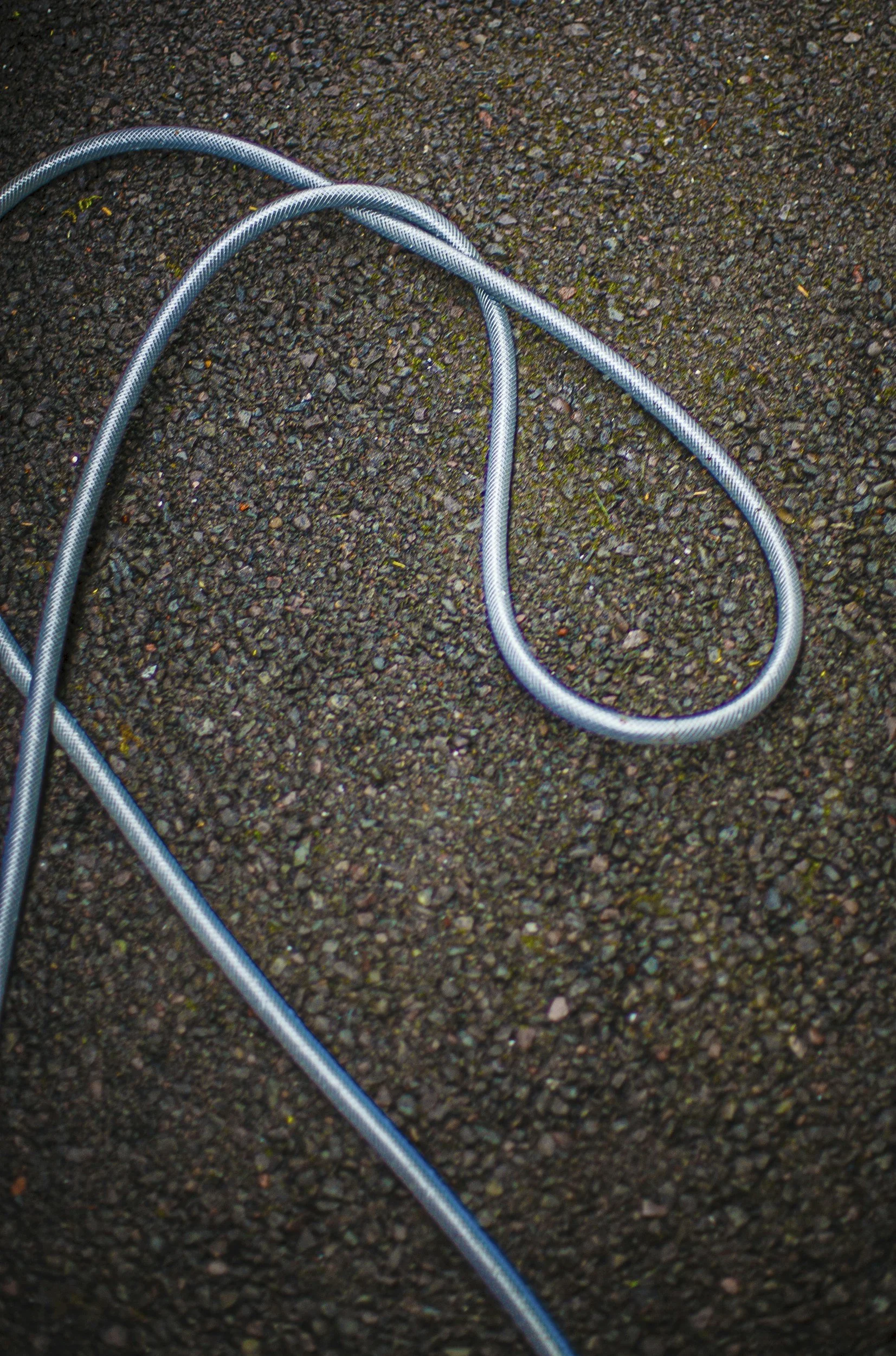A close-up of a tangled silver-gray hose lying on a gravel surface with small stones and patches of moss.