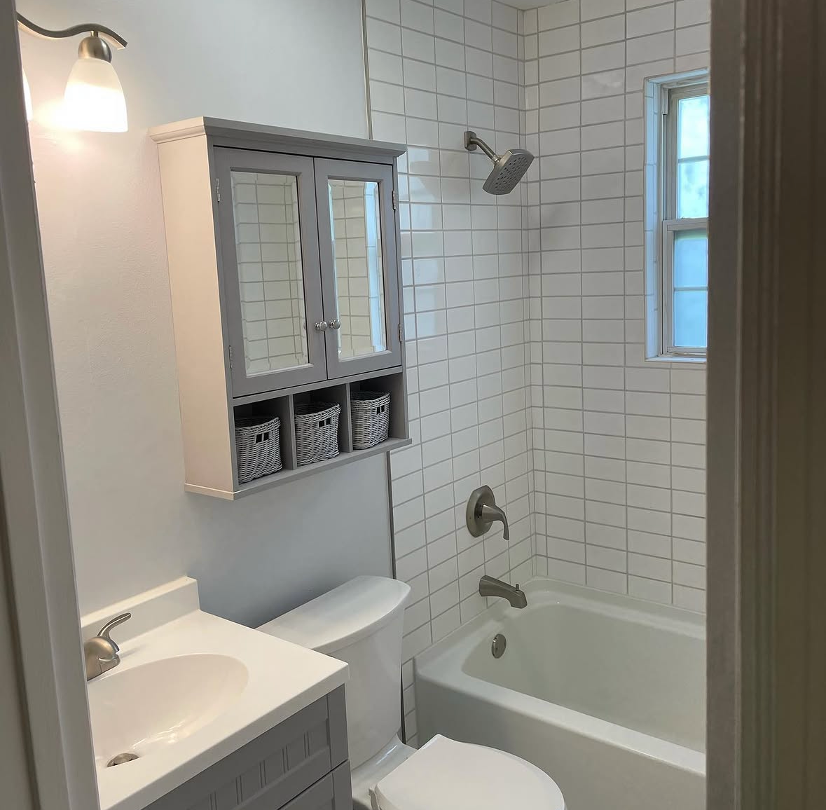 Small bathroom with white tiled walls, a bathtub with a showerhead, a toilet, a small sink with a cabinet, a wall-mounted cupboard with three baskets, a small window, and a wall-mounted light.