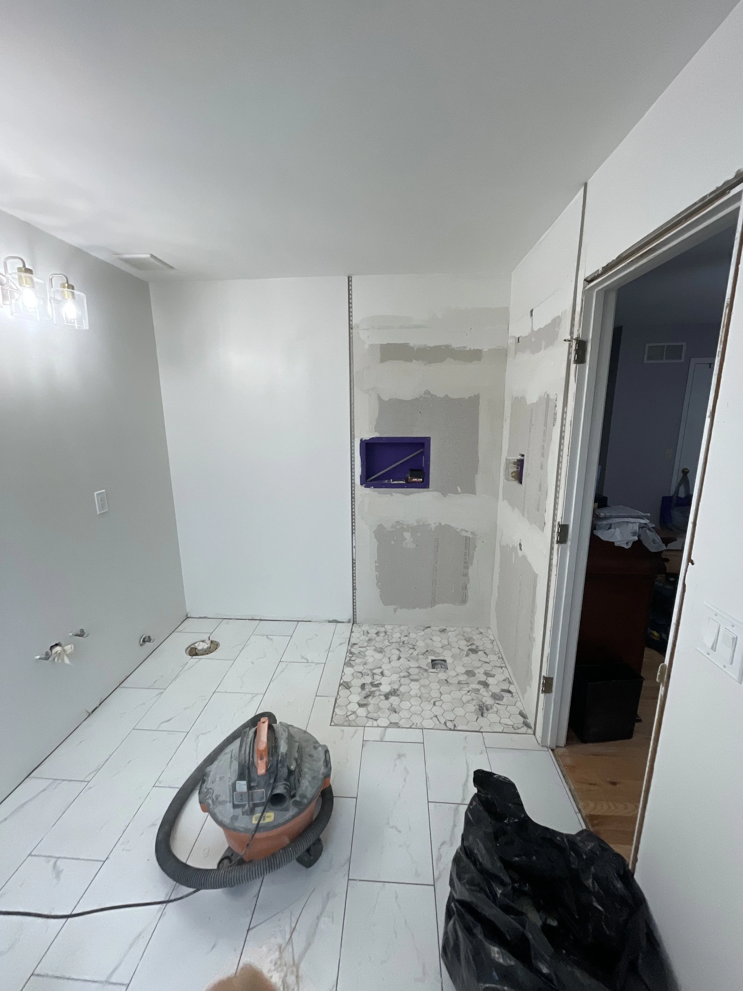 Bathroom under renovation with partially installed white floor tiles, a vacuum cleaner and black trash bags on the floor, and an unfinished wall with a purple electrical box.