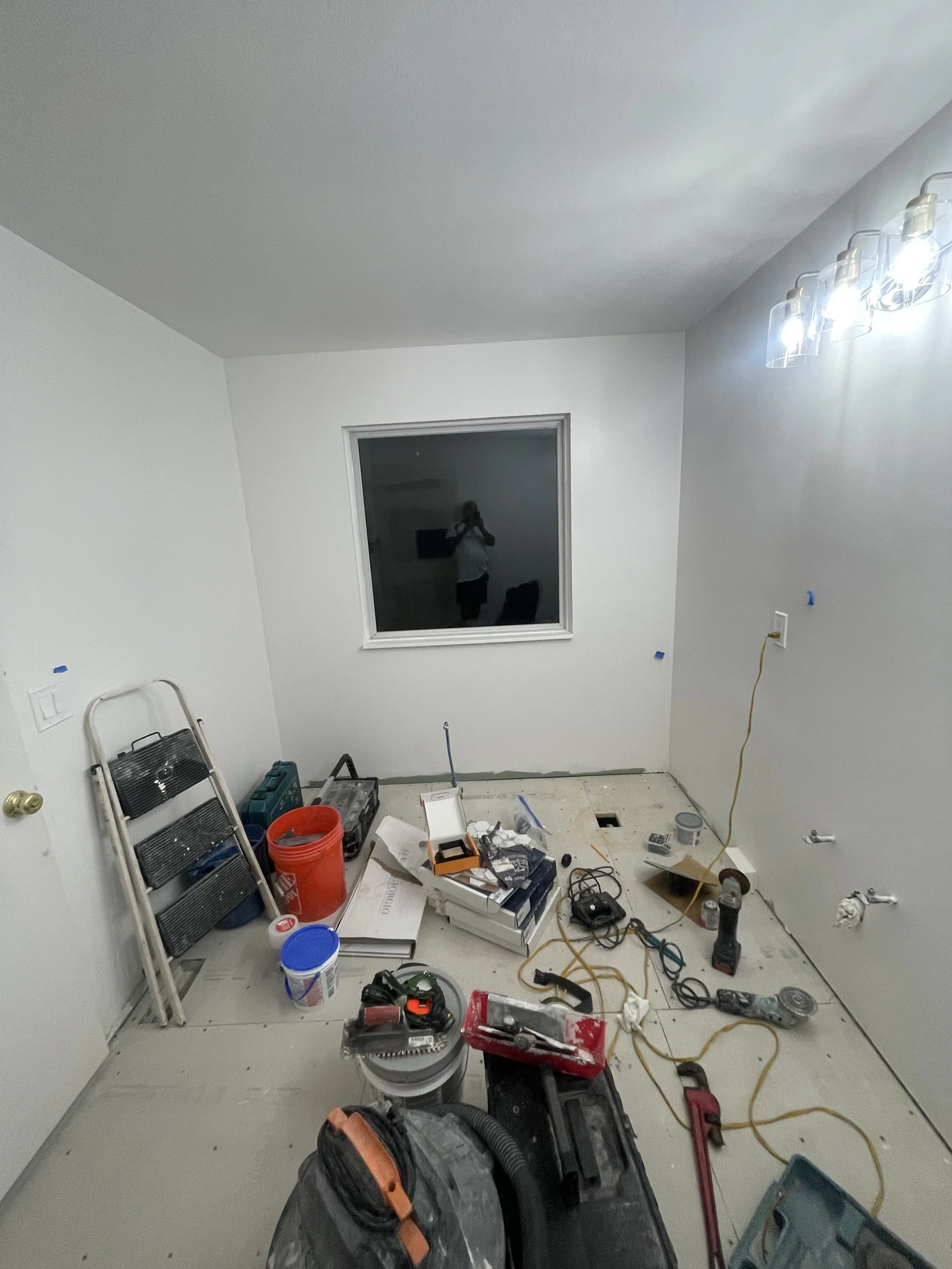 Unfinished room with tools and construction materials scattered on the floor, including a ladder, power tools, paint cans, wires, and a white box with supplies, indicating ongoing renovation or construction work.