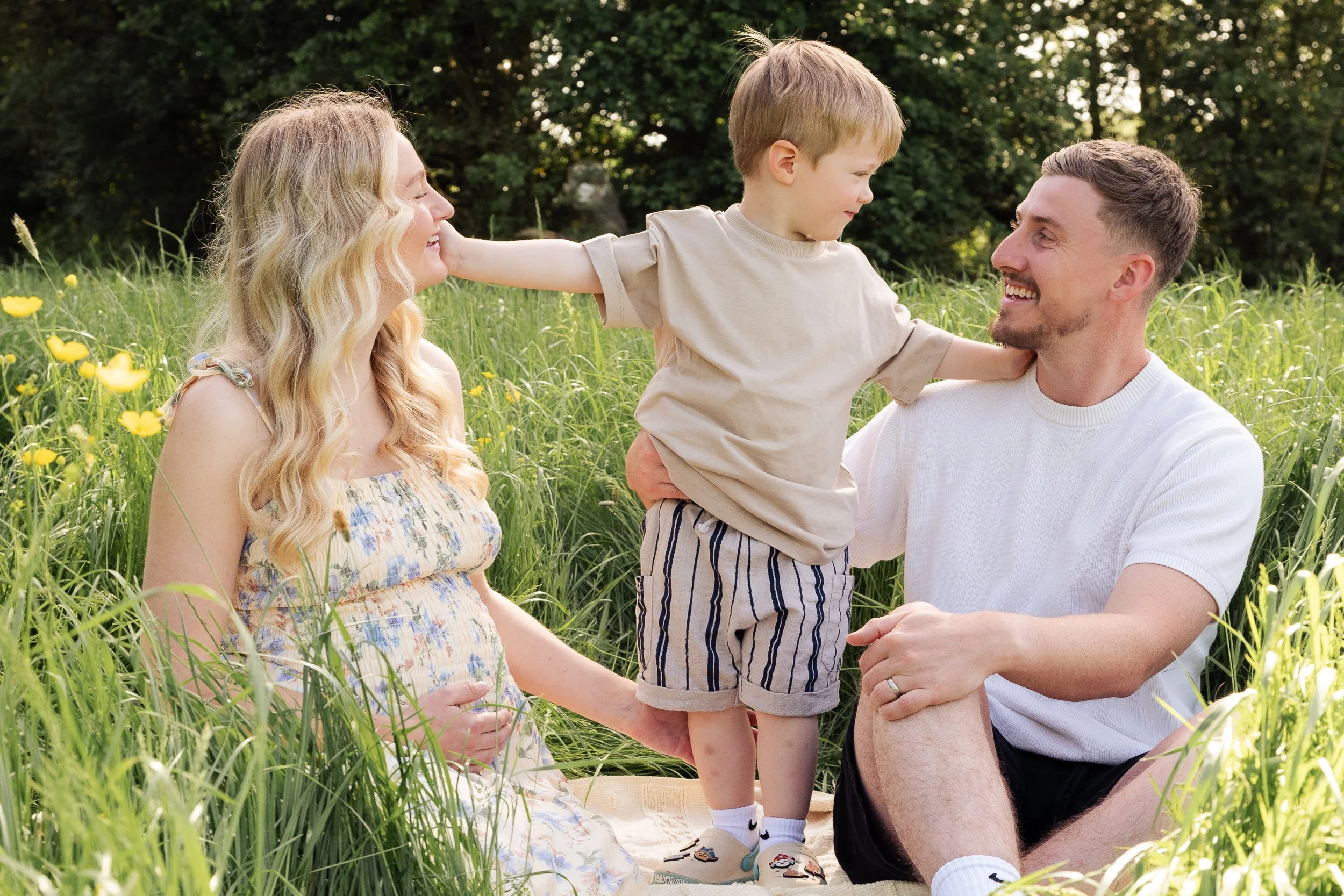 Pregnant mom with toddler and husband sitting on blanket in field of buttercups during a family maternity photoshoot in Worcester, Worcestershire and Droitwich.