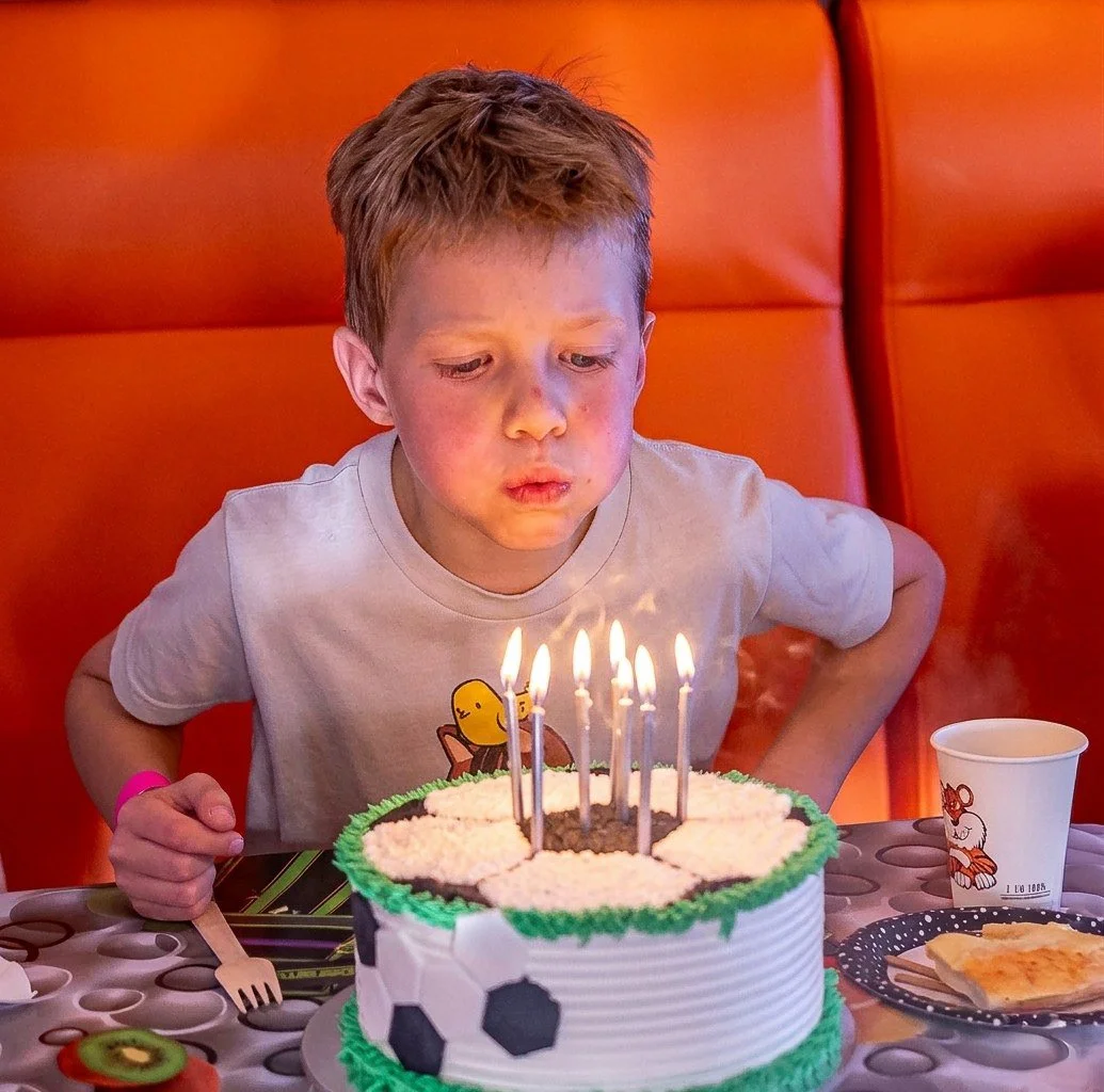 A young boy with red hair blows out candles on a birthday cake decorated with a soccer theme, sitting at a table with party supplies.