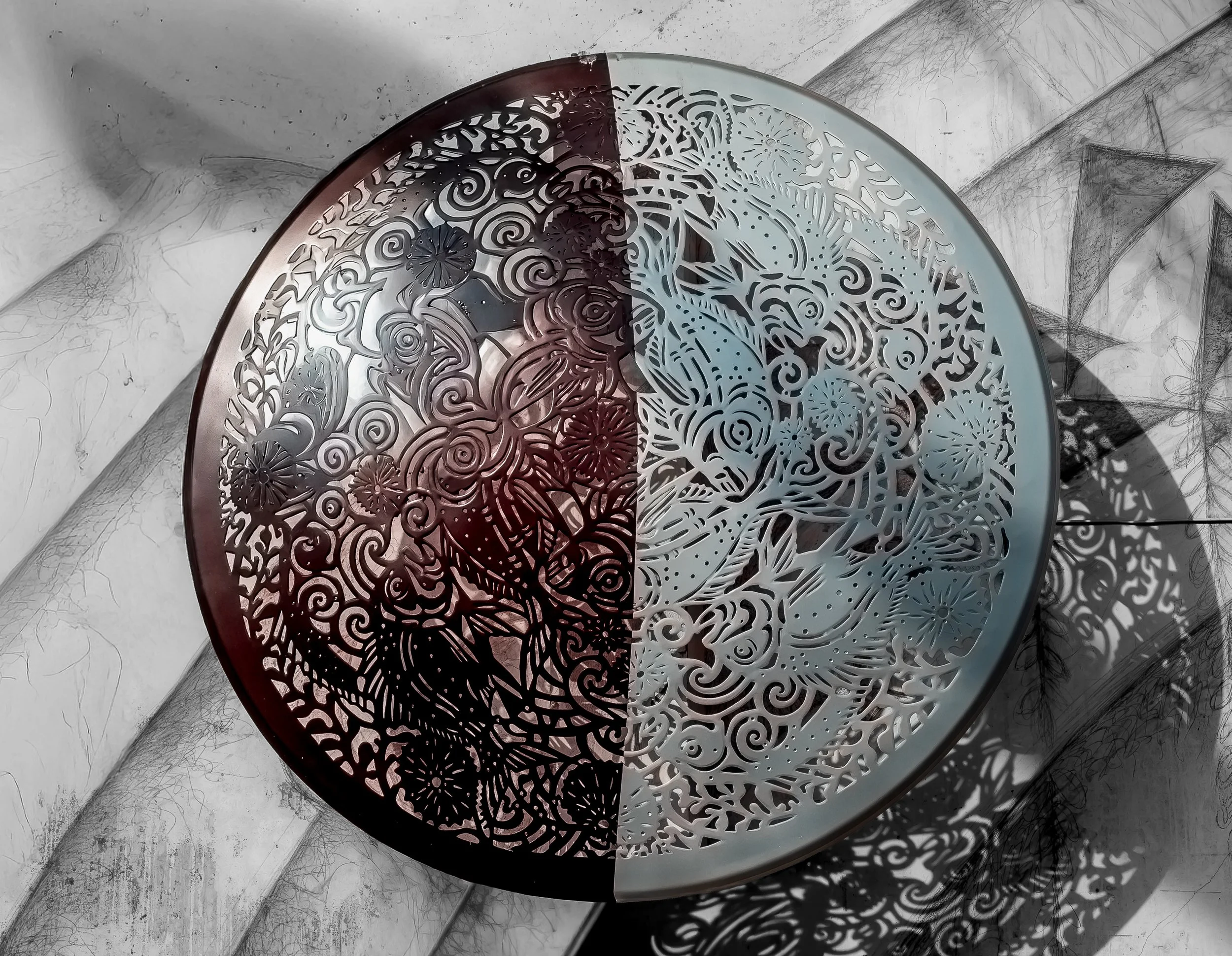 Round metal table with intricate floral and swirl cutout design, two-tone colors, sitting on a concrete floor against a white wall with shadows and cracks.