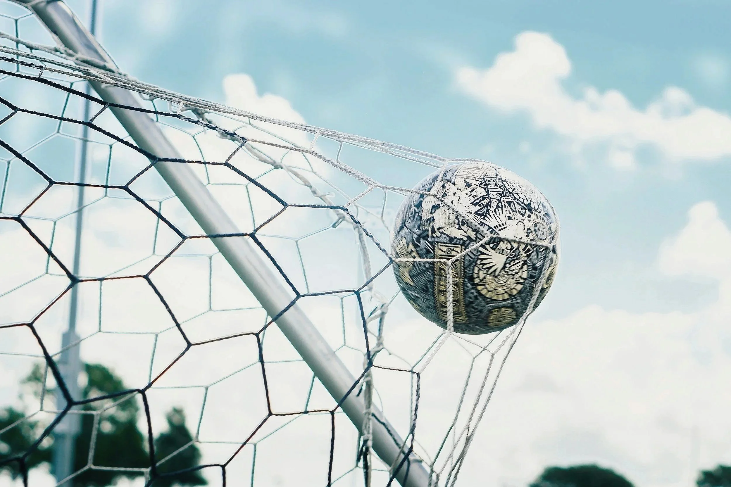 Soccer ball lodged in a soccer goal net against a blue sky with clouds.