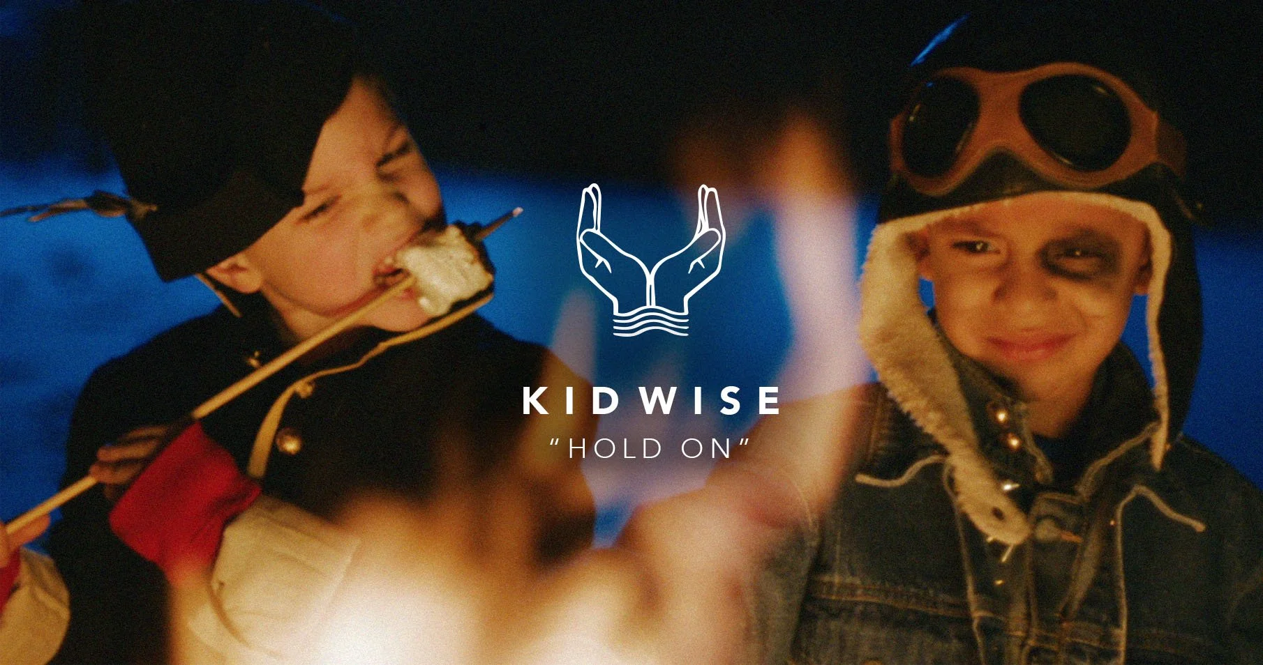 Two young boys wearing aviator helmets and goggles, one smiling with face painted, enjoying a campfire at night.