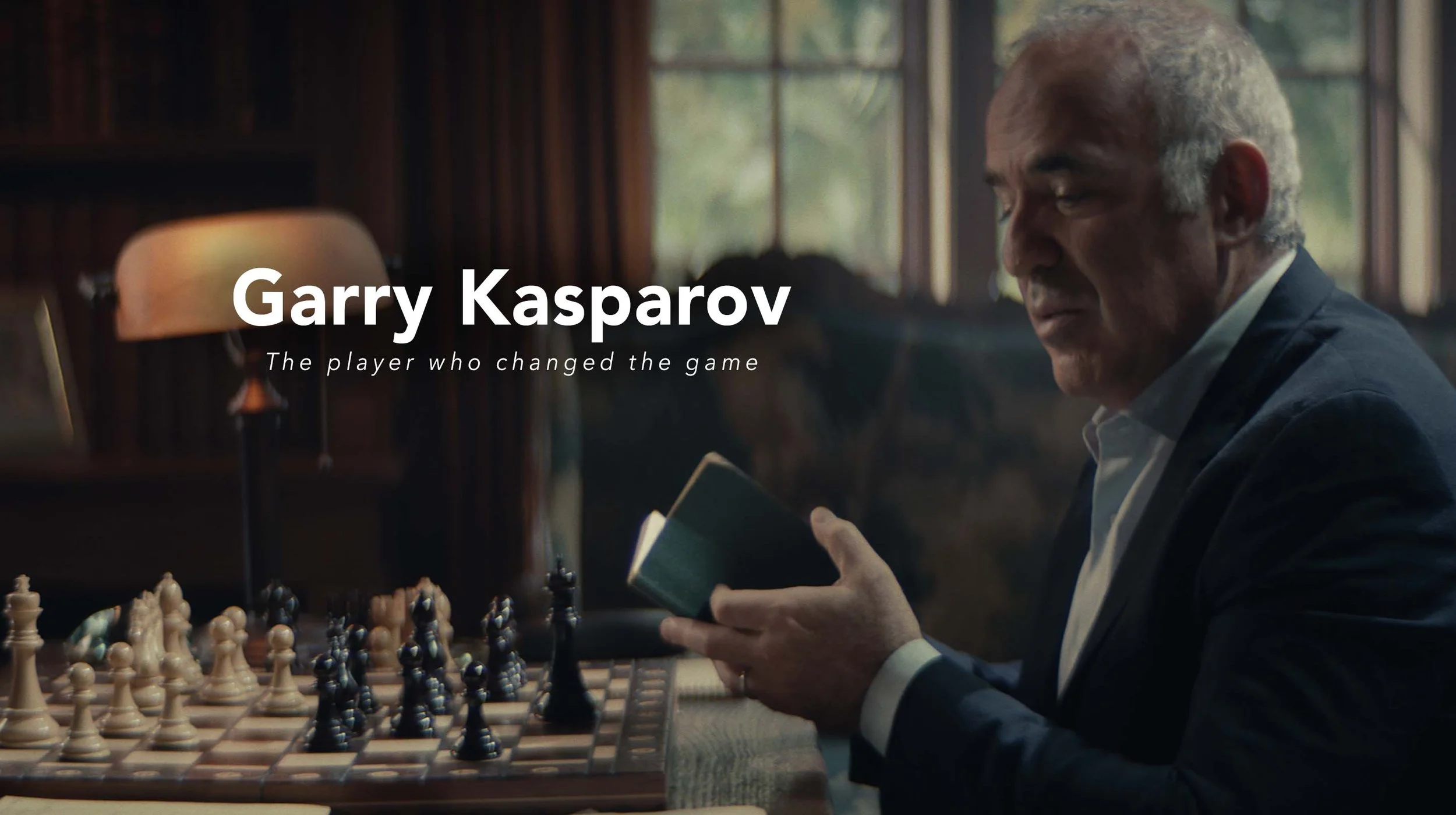 A man reading a book at a chessboard in a cozy, wood-paneled room with large windows, with the text "Garry Kasparov - The player who changed the game" overlaid on the image.