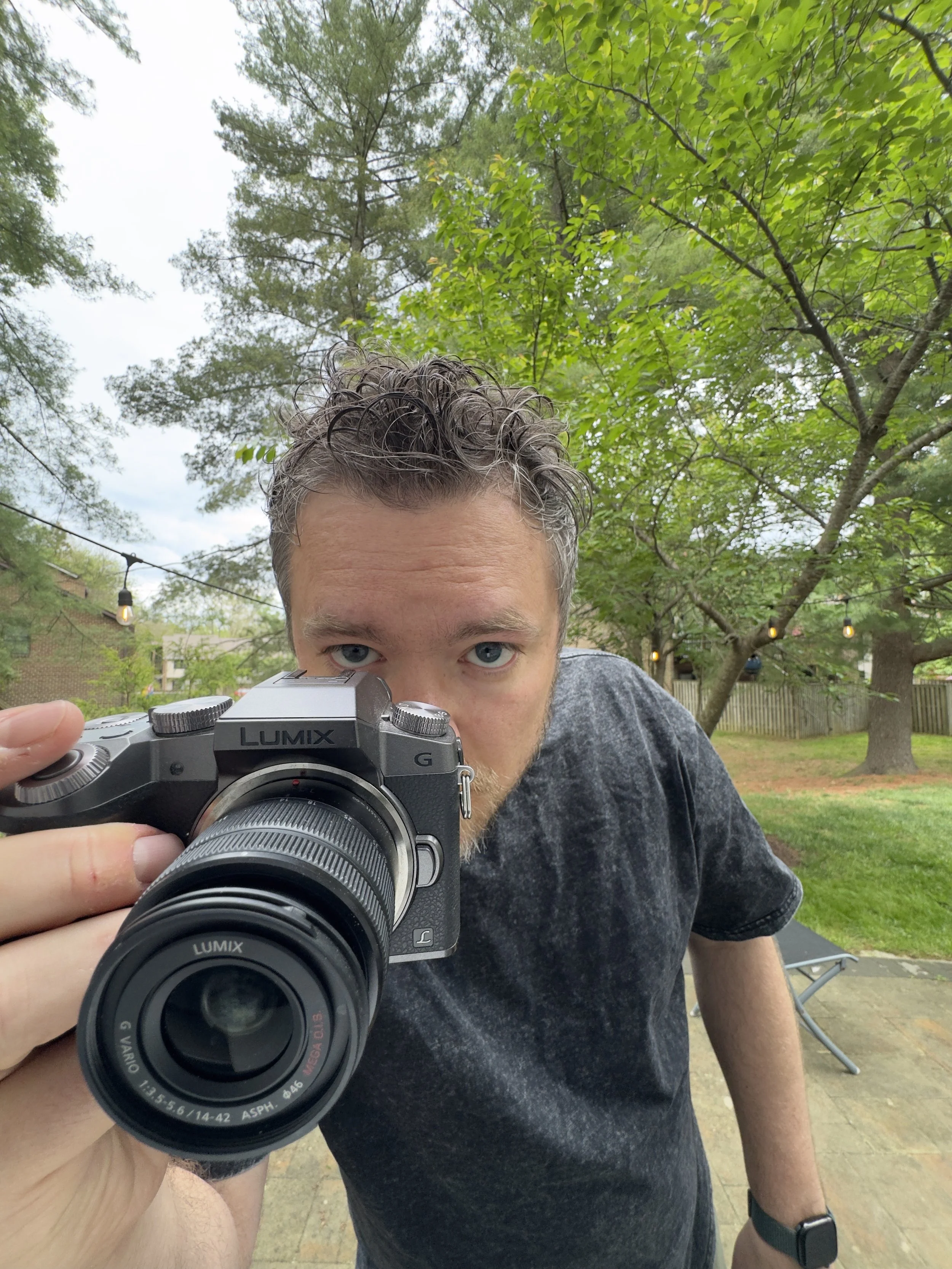 A man with short, curly gray hair and blue eyes is taking a selfie outdoors with a camera, holding the camera with his right hand. He is wearing a dark gray t-shirt, a black wristwatch on his left wrist, and stands in front of green trees and string lights hanging in the background.