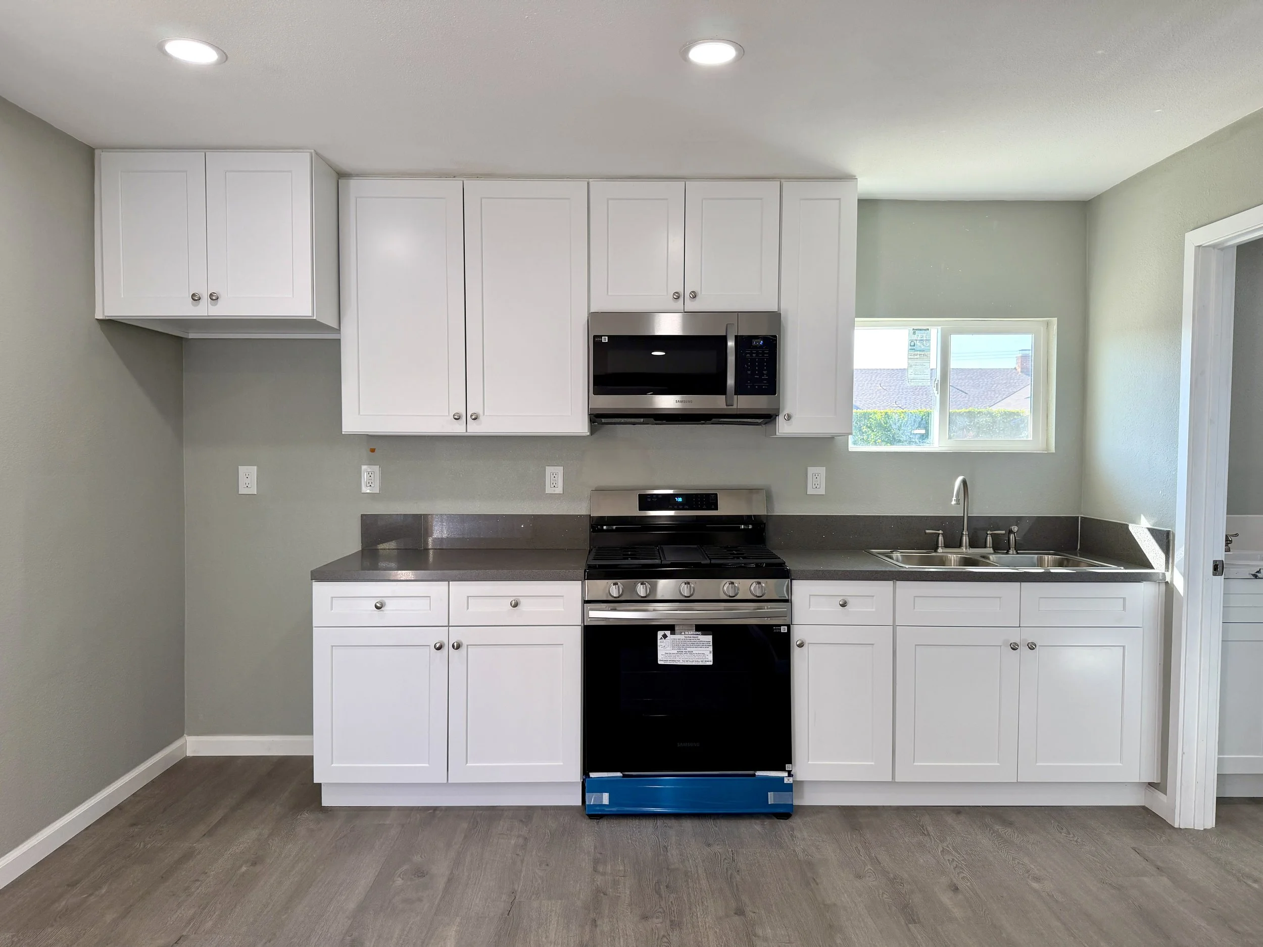 A modern kitchen with white cabinets, stainless steel microwave, oven, and a window letting in natural light. The counter has a stainless steel sink and a small blue object on the floor.