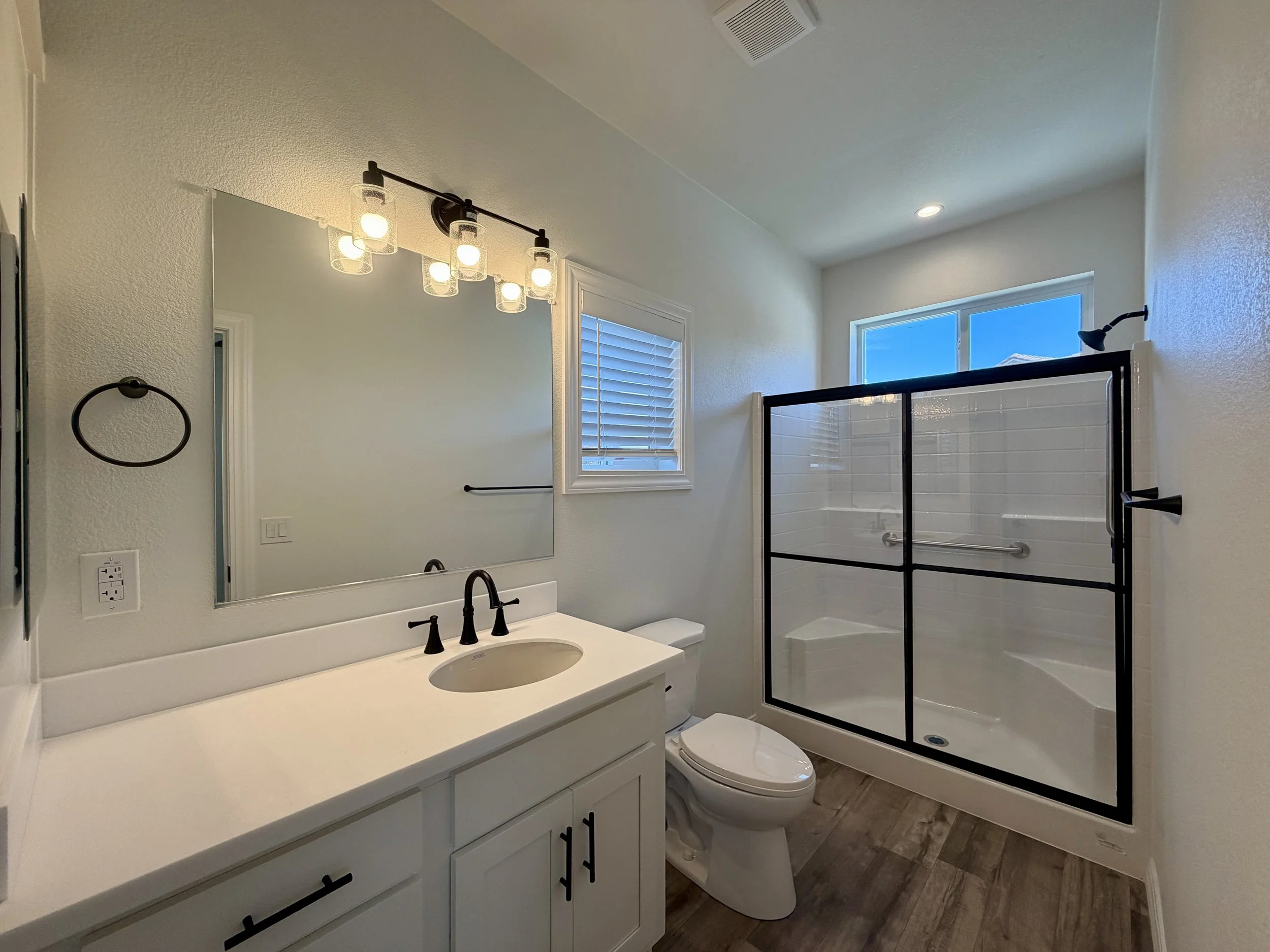 Modern bathroom featuring a white vanity with black hardware, a large mirror, a black faucet, and a white countertop. There is a toilet next to the vanity, a framed glass shower with black hardware, and a window with white shutters. A contemporary light fixture with six bulbs hangs above the mirror, and the ceiling has a vent and recessed lighting.