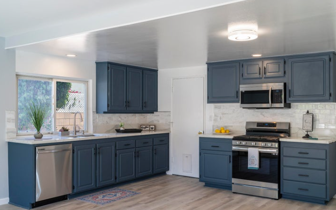 Kitchen with blue cabinets, stainless steel stove and microwave, white backsplash, window with view outside, decor items, and wooden flooring.