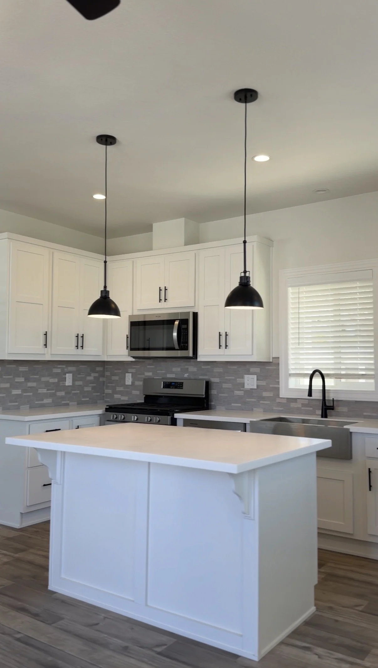 Modern kitchen with white cabinets, a gray tile backsplash, black appliances, black pendant lights, a white kitchen island, and a window with white blinds.