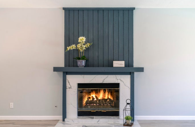 Living room fireplace with blue wooden panel above mantel, decorated with a vase of white flowers and a small sign, with a modern fireplace, marble surround, and a lantern with plants on the floor.