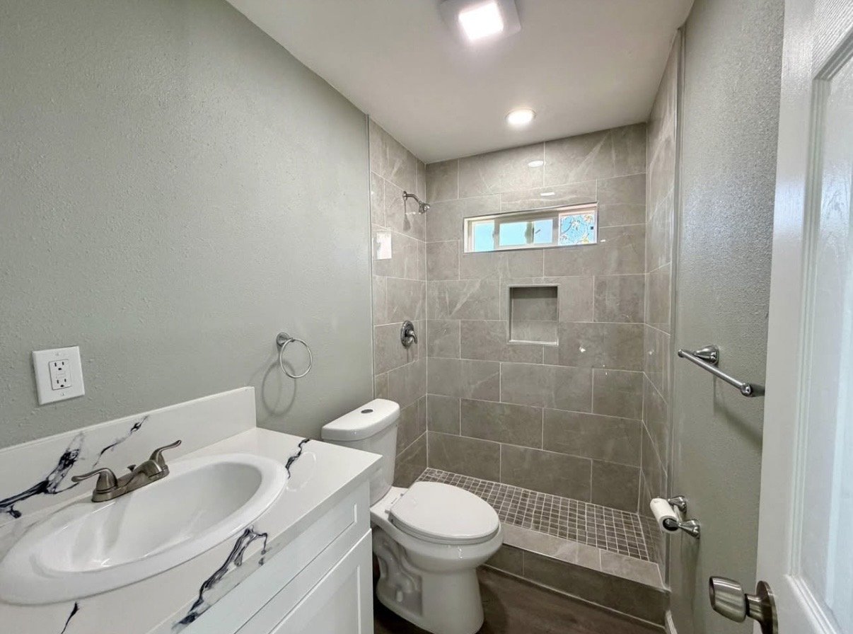 Bathroom with a marble-top vanity, sink, toilet, and walk-in shower with beige tiles, a small high window, and ceiling lights.