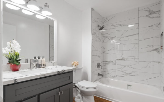 Modern bathroom with a white marble shower and bathtub, a gray vanity with a marble countertop, a mirror with lights, and decorative items.