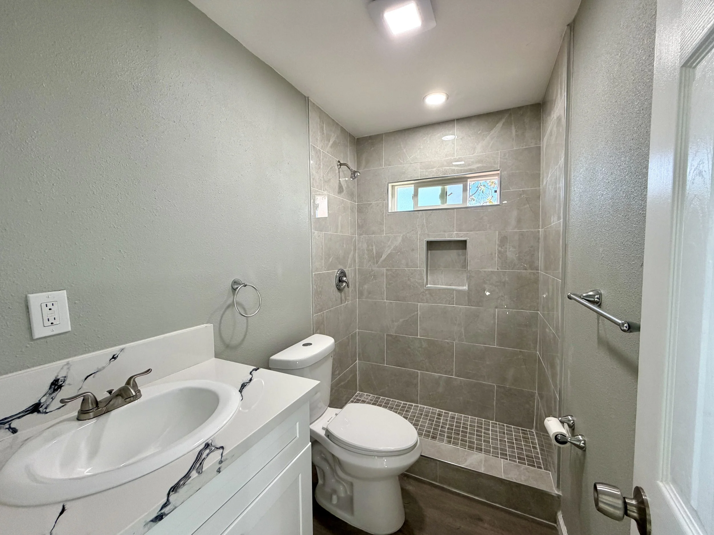 Bathroom with a white vanity and sink, a toilet, a shower with beige tiles, and a small window.
