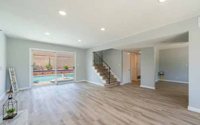 Empty living room with light wood flooring, white walls, sliding glass door to backyard, and staircase with dark handrail.