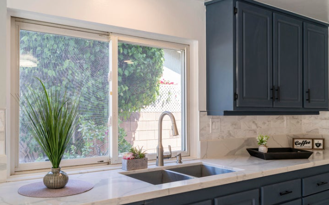 Kitchen window with blue cabinets, white countertop, a sink, green plant, and outside view of trees and fence.