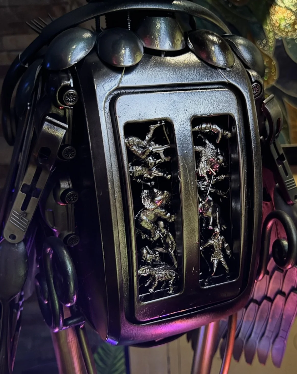 A close-up of a vintage metal fire alarm box filled with small metallic action figures of soldiers and animals, surrounded by various metal tools and decorative elements.