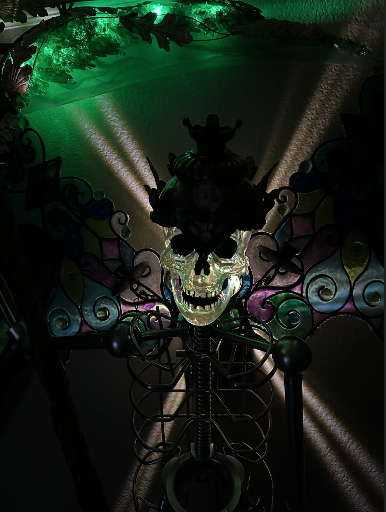 A skull-shaped decorative object illuminated with colorful lights, placed on a black metal stand, viewed through a decorative metal and glass frame with colored accents. The scene is dimly lit, creating a spooky atmosphere with green, purple, and yellow hues on the textured wall in the background.
