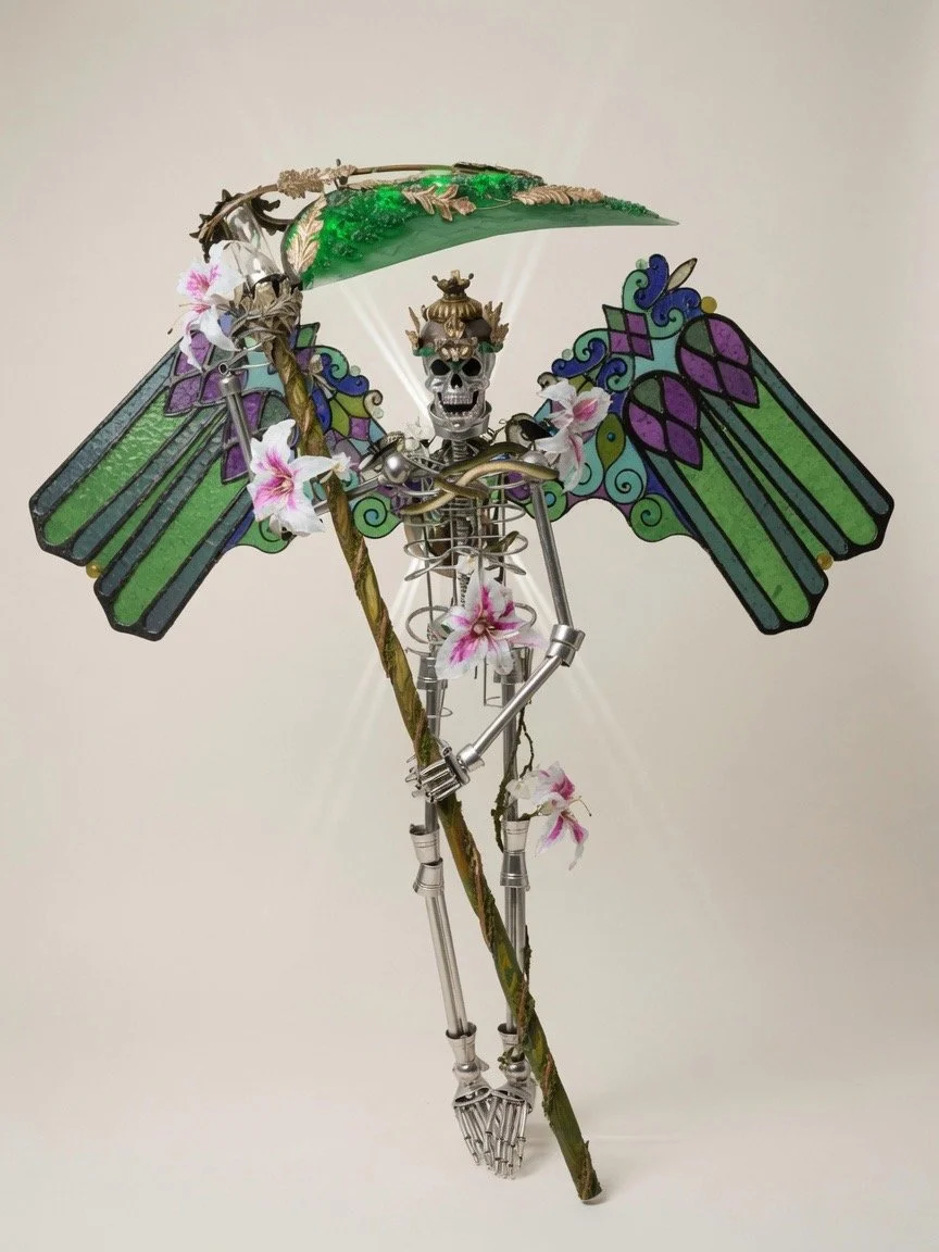 A metal skeleton figure with dragonfly-like wings, holding a decorative staff topped with a shade and faux flowers, standing against a plain background.