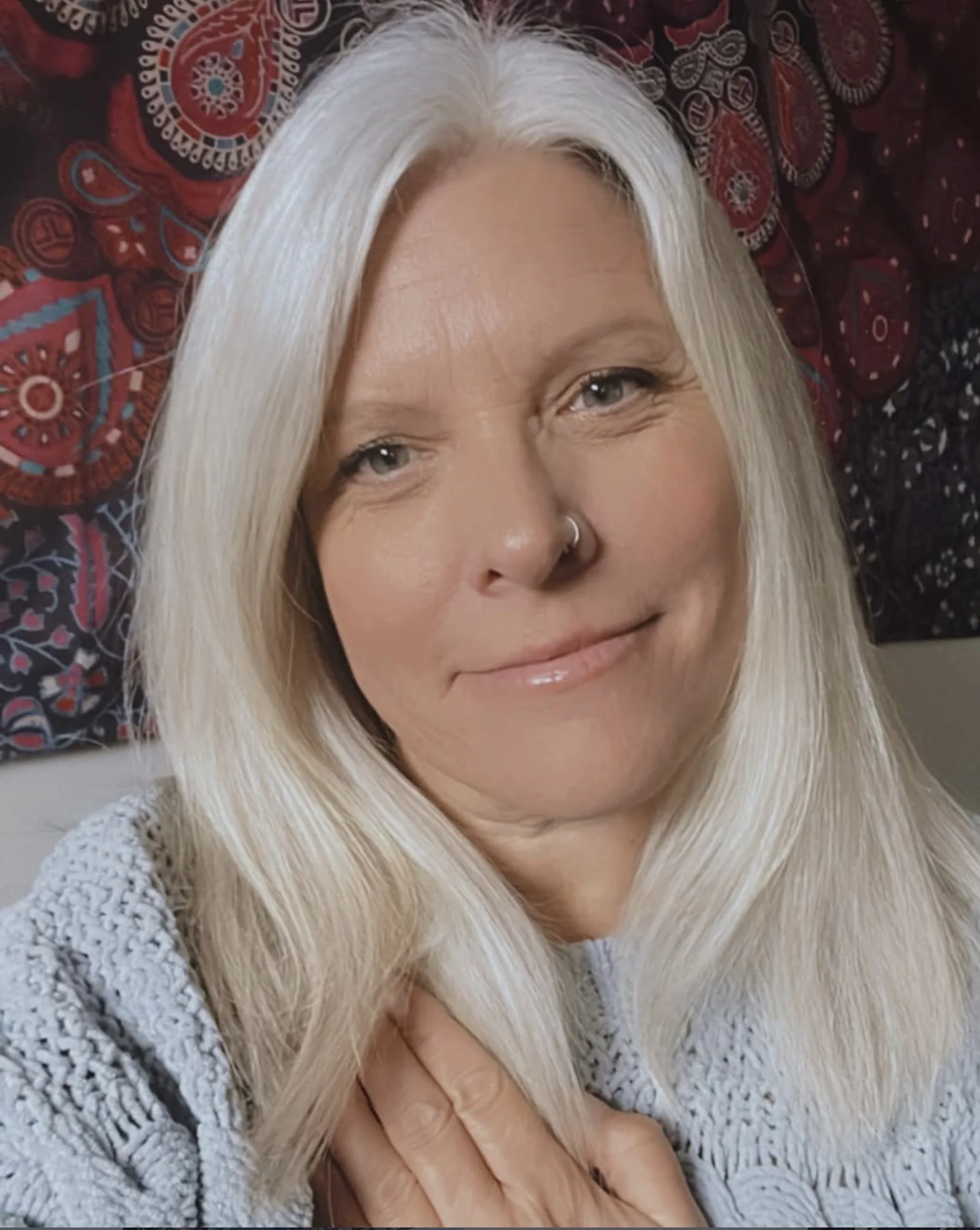 A woman with long, straight platinum blonde hair, blue eyes, and a nose ring, smiling while laying down on a patterned blanket and wearing a gray knit sweater.