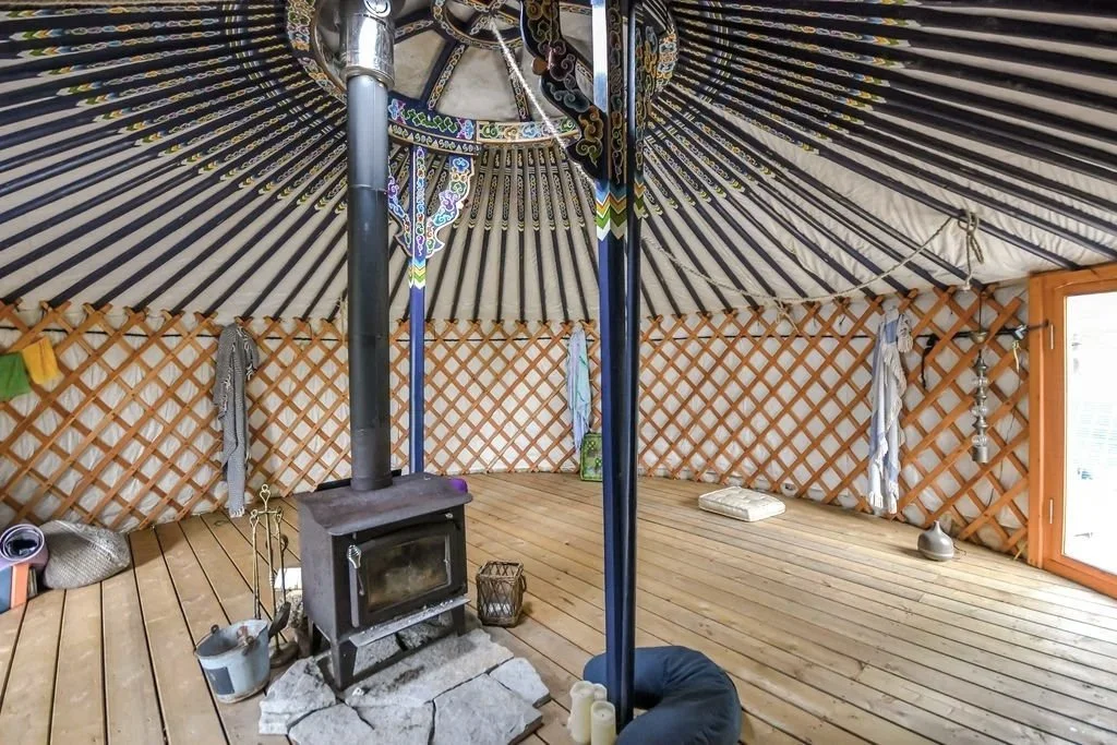 Interior of a yurt with wooden flooring, a wood stove, and decorative textiles hanging on the lattice walls.
