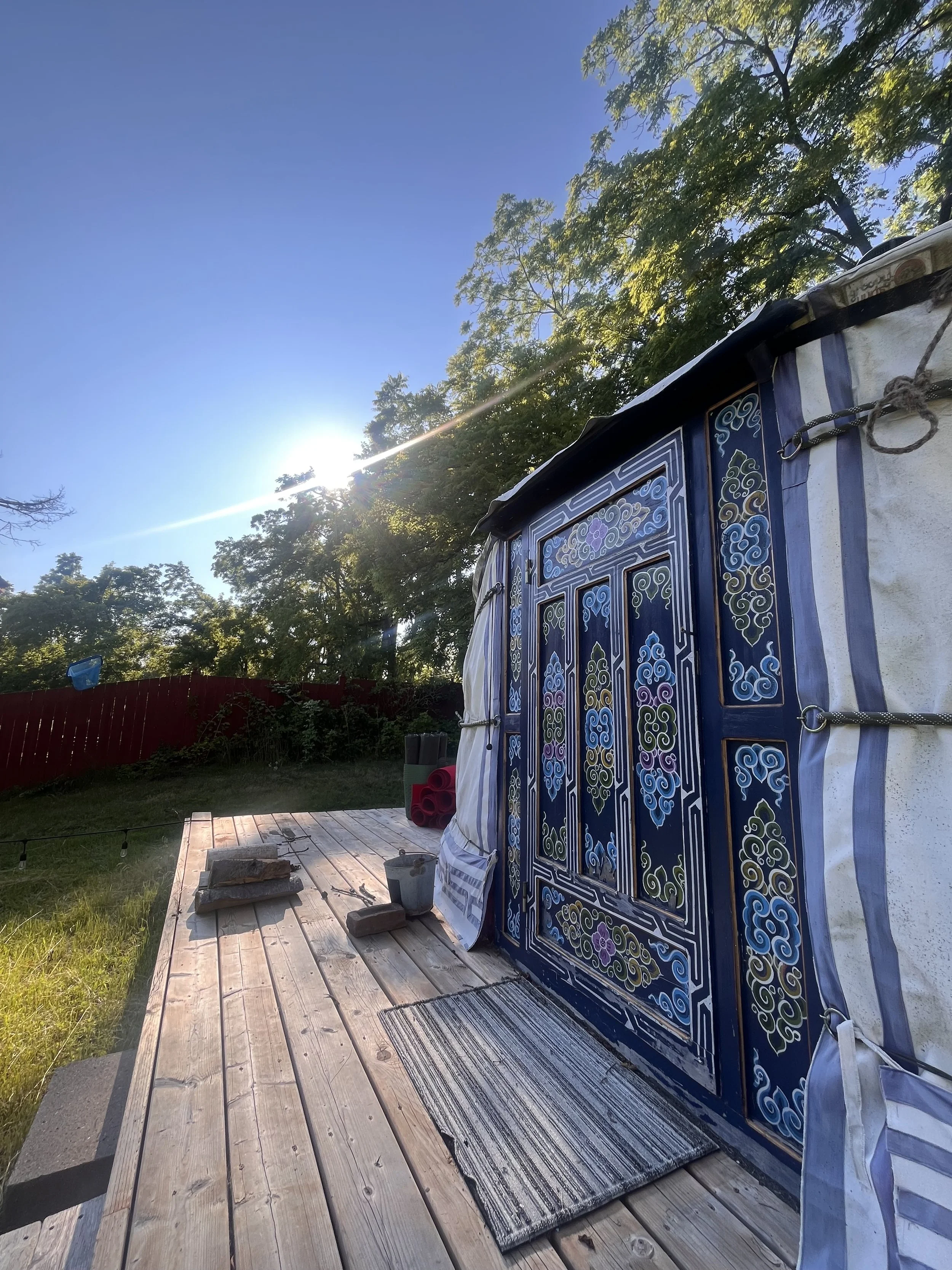 A tent with intricate blue, gold, and purple patterns set up on a wooden platform in a backyard, with green trees, a red fence, and the sun shining in a clear blue sky in the background.