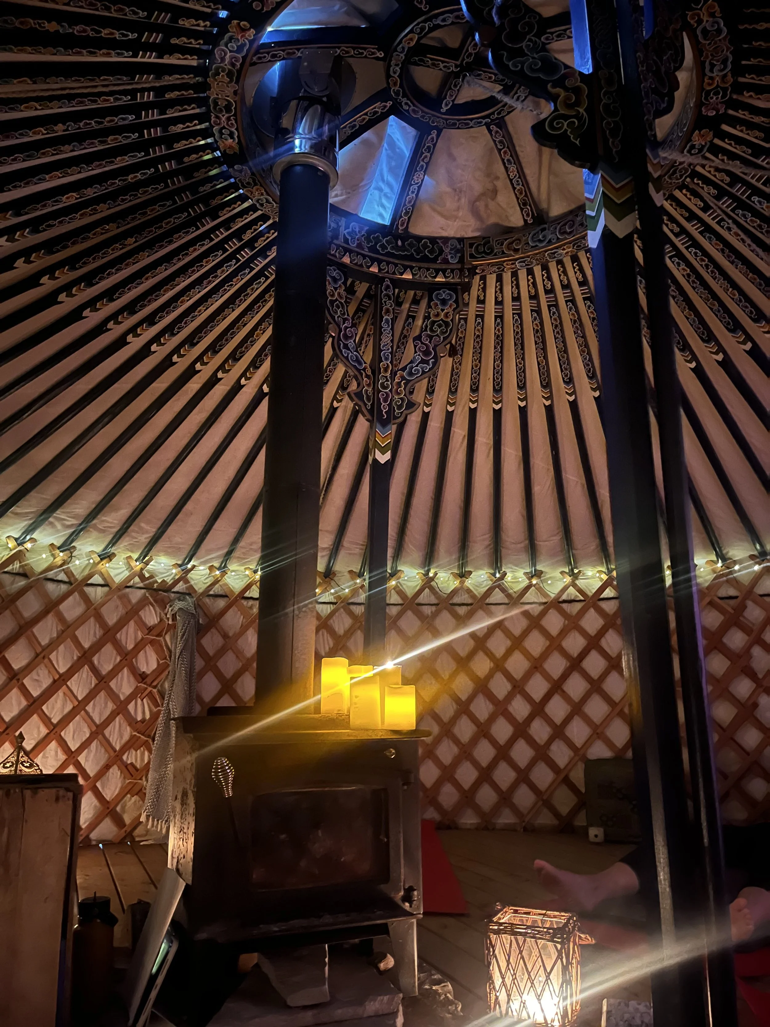 Inside a yurt with a wooden structure, decorative embroidery, a central wooden pole, a small black stove, yellow candles, and a red rug on the floor.