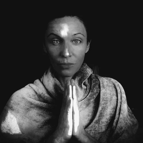 A woman with a serious expression, wearing a gray scarf, with her hands pressed together in a prayer or meditative pose, against a black background.