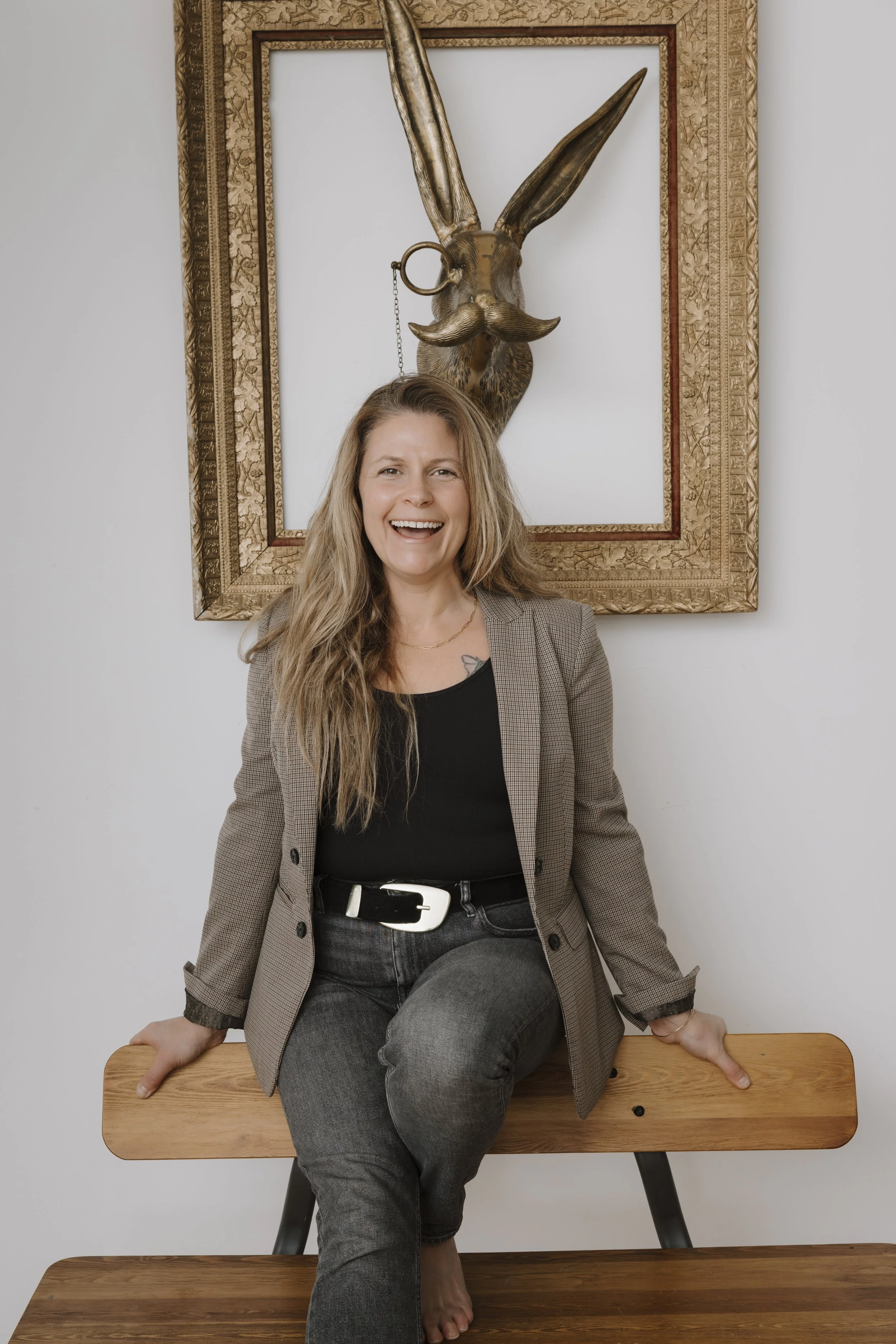 A smiling woman with long hair, standing on a wooden bench, wearing a gray checkered blazer, black shirt, and gray pants, with her right hand resting on the bench and her left hand on her knee. She is seated in front of a wall with a framed piece of art featuring a metallic rabbit head with a mustache and a ring in one of its ears.