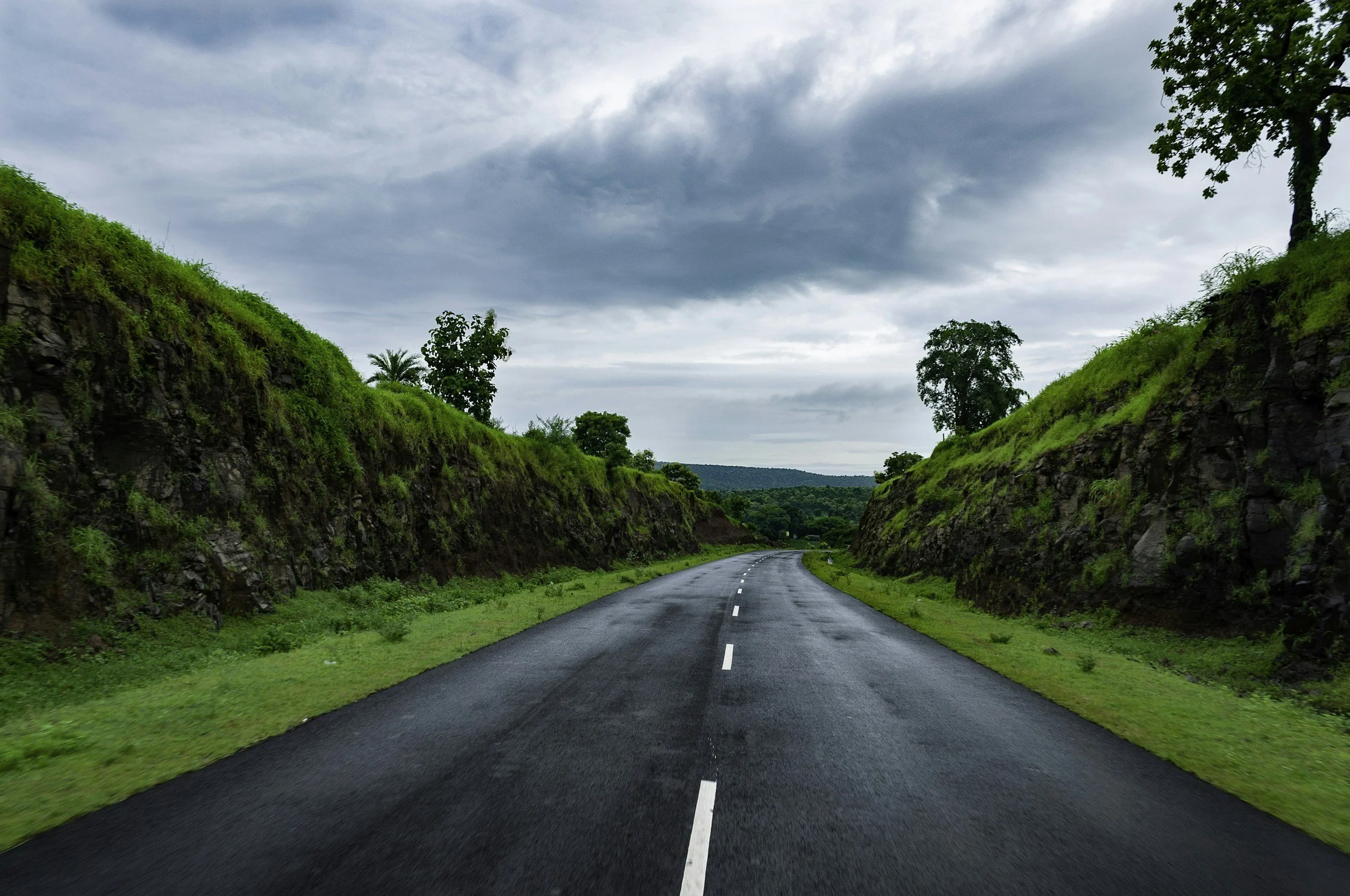 A paved road curving through a green landscape with large rocks on the sides and trees in the background, under a cloudy sky.