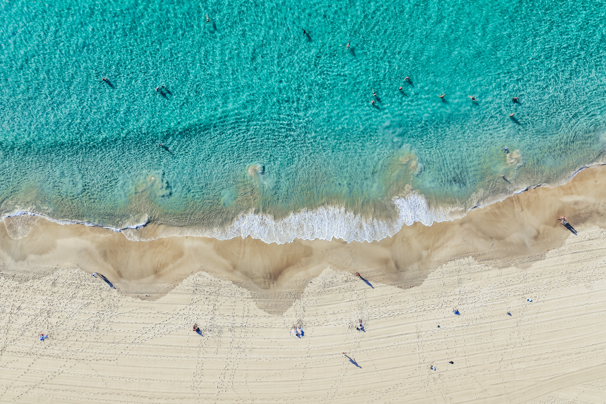 Aerial view of a beach with light-colored sand, turquoise waves, and several people walking or sitting on the shore.