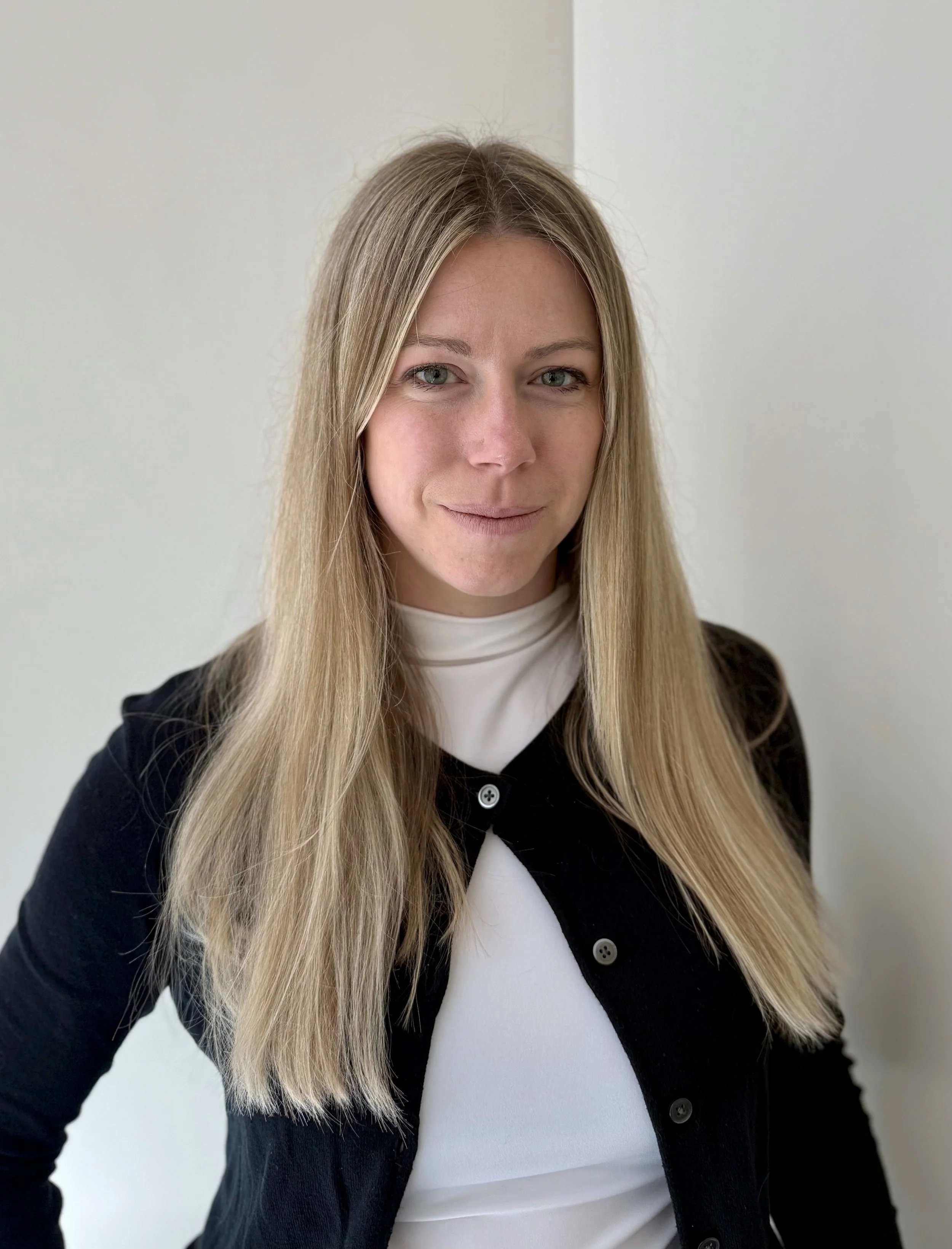 Close-up portrait of a woman with long blonde hair, wearing a white top and black cardigan, standing against a white background.